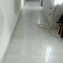 REM-BUD PIOTR SIKORA - Jasne, błyszczące płytki podłogowe ułożone w korytarzu, widoczne ślady prac na ścianie. W tle sylwetka osoby. Oświetlenie budowlane po prawej.