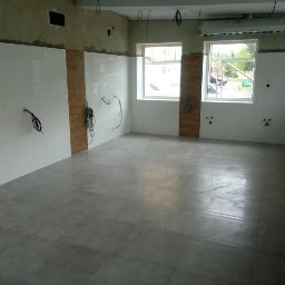 REM-BUD PIOTR SIKORA - Wnętrze w trakcie prac wykończeniowych: ułożona szara podłoga z płytek, częściowo wykończone ściany z białymi płytkami, widoczne kable elektryczne.