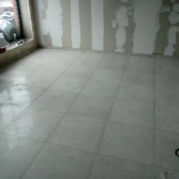 REM-BUD PIOTR SIKORA - Wykończona podłoga z jasnych, kwadratowych płytek w nowym pomieszczeniu. Na ścianach widoczne ślady po szpachlowaniu, okno z lewej strony. Kabel na podłodze.