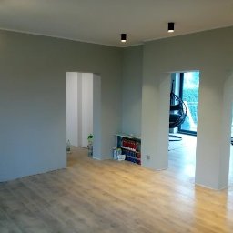 REM-BUD PIOTR SIKORA - Jasne, przestronne wnętrze po remoncie: świeżo pomalowane ściany w odcieniach szarości, ułożone panele podłogowe, widoczne oświetlenie sufitowe i przejścia do innych pomieszczeń.