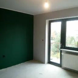 Pokój po malowaniu: ściana w odcieniu butelkowej zieleni, szara ściana z oknem balkonowym. Widoczny grzejnik i surowa podłoga.