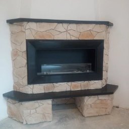 REM-BUD PIOTR SIKORA - Kanciasty kominek obłożony jasnym kamieniem dekoracyjnym, z czarnym wkładem i blatem, umieszczony w rogu pomieszczenia. Wykończenie wnętrza.