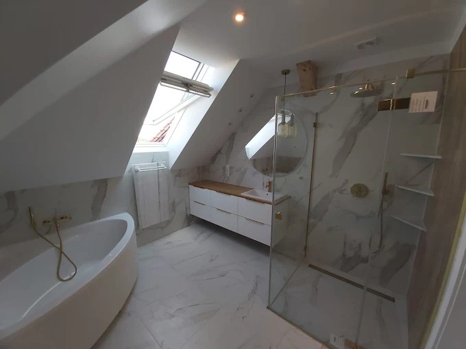 Jasna łazienka na poddaszu z wanną, kabiną prysznicową i umywalką. Białe meble i marmurowe płytki tworzą elegancki wystrój. Okno dachowe doświetla wnętrze.