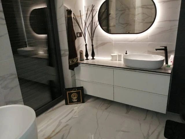 Nowoczesna łazienka z owalnym lustrem podświetlanym LED, białą umywalką nablatową i szafką. Marmurowe płytki na ścianach i podłodze. Elegancki design.