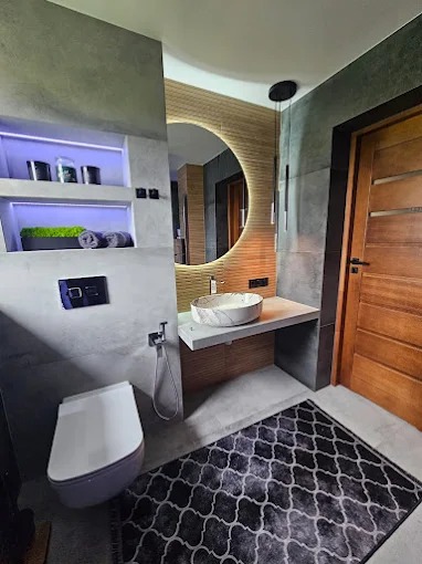 Nowoczesna łazienka z okrągłym lustrem z podświetleniem LED, marmurową umywalką i betonową zabudową. Szara armatura i dywan w marokański wzór dopełniają wystrój.