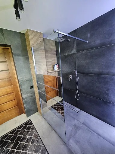 Nowoczesna łazienka z kabiną typu walk-in, szare płytki imitujące beton, drewniane akcenty i designerska lampa sufitowa. Widoczny odpływ liniowy i minimalistyczna armatura.