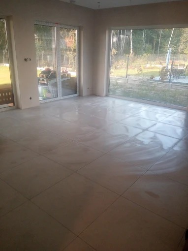 Jasne, świeżo położone płytki podłogowe w przestronnym salonie z dużymi oknami wychodzącymi na ogród. Widoczne niedokończone ściany, przygotowane do malowania.