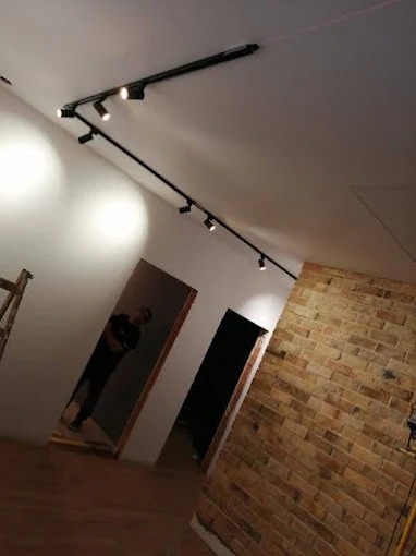 Nowoczesne oświetlenie szynowe z reflektorami w świeżo wykończonym wnętrzu z ceglaną ścianą i drewnianą podłogą. Widoczna osoba w tle.