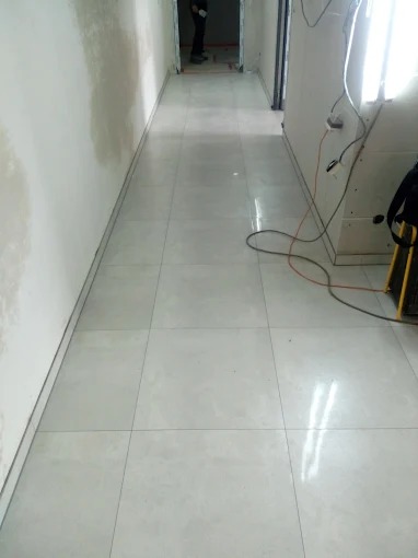 Jasne, błyszczące płytki podłogowe ułożone w korytarzu, widoczne ślady prac na ścianie. W tle sylwetka osoby. Oświetlenie budowlane po prawej.