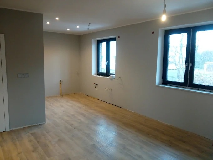 Jasne panele podłogowe w przestronnym wnętrzu po remoncie. Ściany pomalowane na szaro, widoczne dwa okna z czarnymi ramami. Oświetlenie sufitowe.