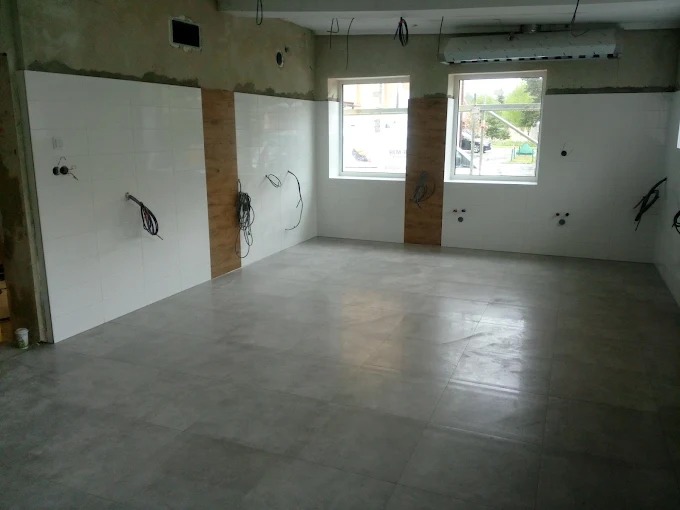 Wnętrze w trakcie prac wykończeniowych: ułożona szara podłoga z płytek, częściowo wykończone ściany z białymi płytkami, widoczne kable elektryczne.