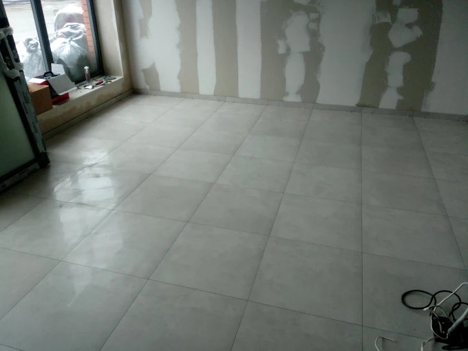 Wykończona podłoga z jasnych, kwadratowych płytek w nowym pomieszczeniu. Na ścianach widoczne ślady po szpachlowaniu, okno z lewej strony. Kabel na podłodze.