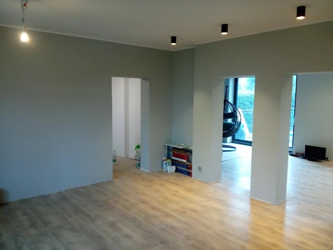 Jasne, przestronne wnętrze po remoncie: świeżo pomalowane ściany w odcieniach szarości, ułożone panele podłogowe, widoczne oświetlenie sufitowe i przejścia do innych pomieszczeń.