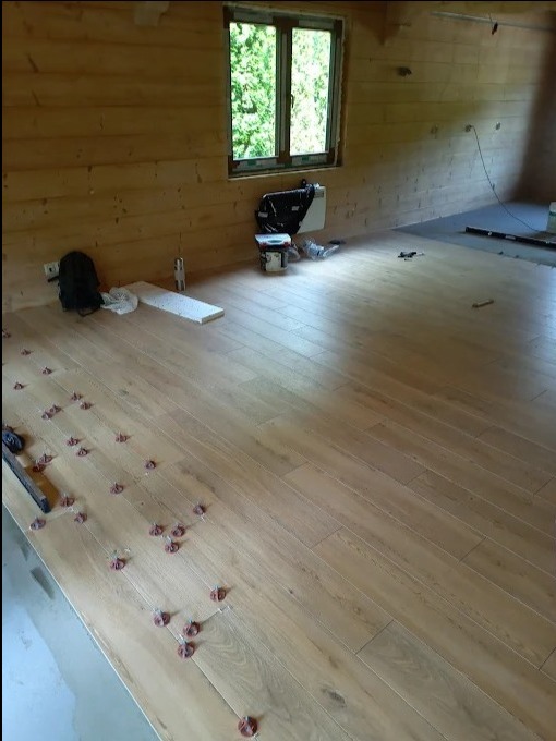 Jasne panele podłogowe w trakcie montażu w drewnianym wnętrzu. Widoczne narzędzia i elementy montażowe, okno z widokiem na zieleń. Częściowo ułożona podłoga.