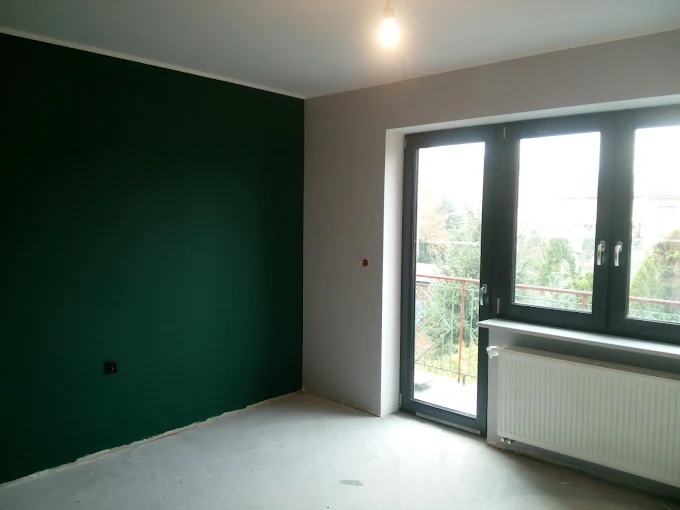 Pokój po malowaniu: ściana w odcieniu butelkowej zieleni, szara ściana z oknem balkonowym. Widoczny grzejnik i surowa podłoga.