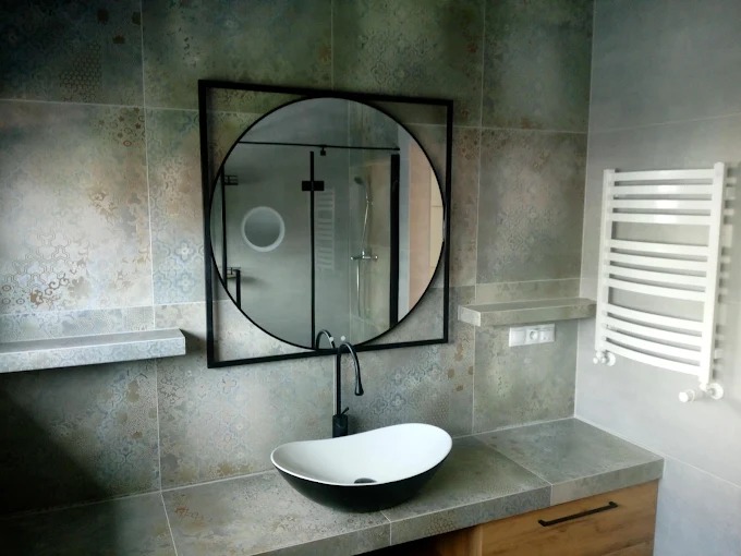 Wykończona łazienka z umywalką wpuszczaną w blat, okrągłym lustrem w czarnej ramie i grzejnikiem drabinkowym. Na ścianach wzorzyste płytki w odcieniach szarości i beżu.