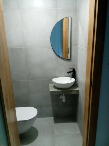 Wykończona łazienka w szarościach z umywalką nablatową, półokrągłym lustrem i podwieszaną toaletą. Minimalistyczny design, płytki wielkoformatowe, czarna armatura.