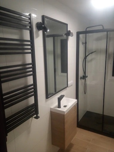 Nowoczesna łazienka z czarnymi akcentami: kabina prysznicowa, grzejnik drabinkowy, lustro z oświetleniem i szafka z umywalką w jasnym drewnie.