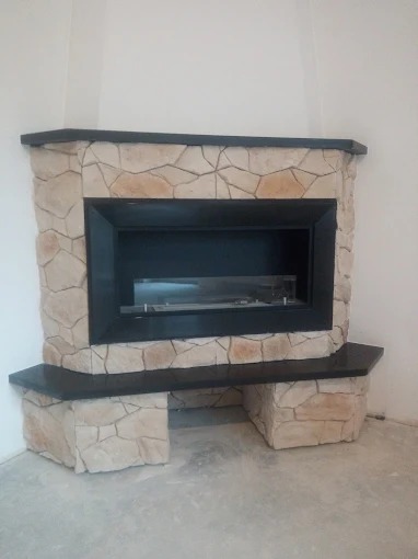 Kanciasty kominek obłożony jasnym kamieniem dekoracyjnym, z czarnym wkładem i blatem, umieszczony w rogu pomieszczenia. Wykończenie wnętrza.