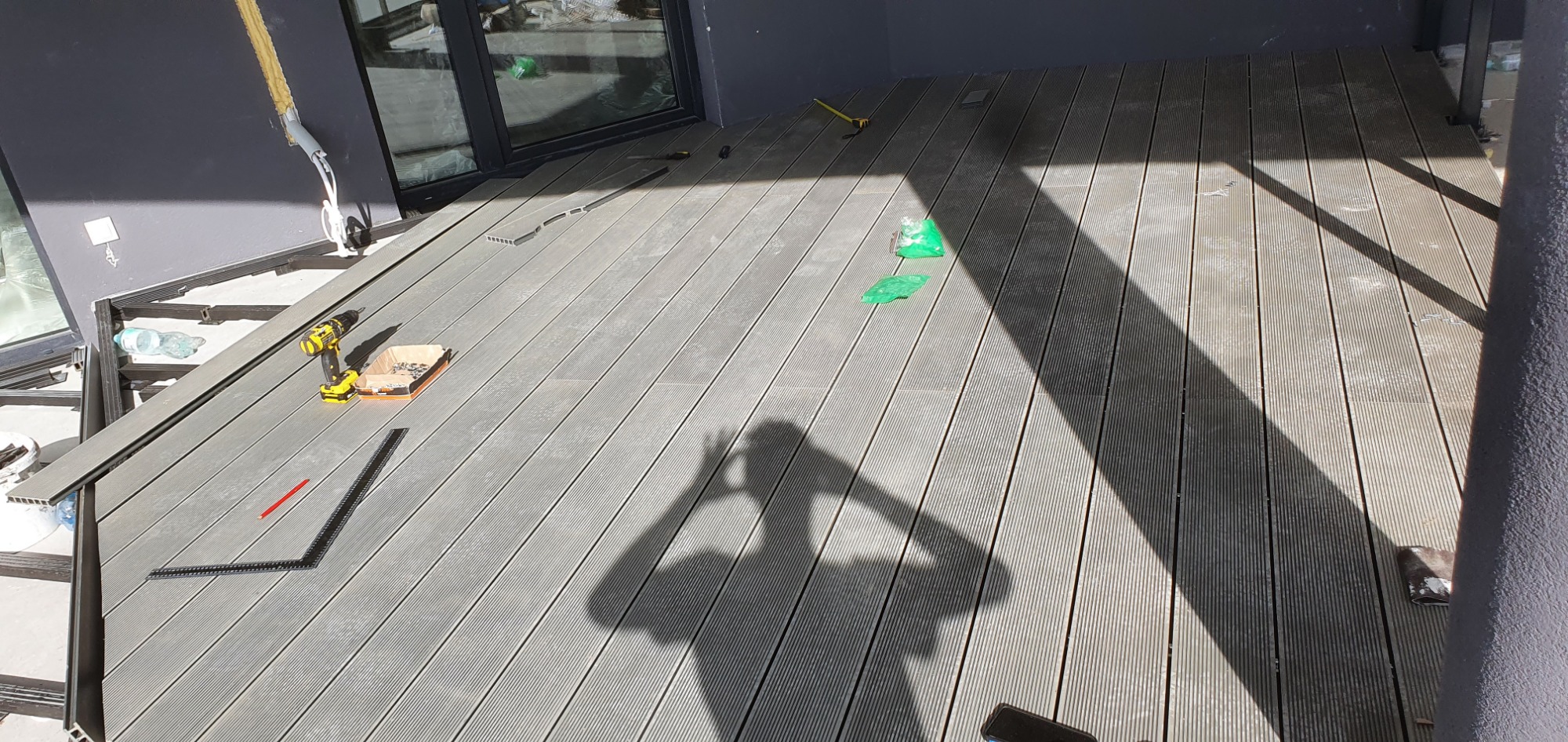 Montaż szarego tarasu kompozytowego na balkonie z widocznymi narzędziami: wkrętarka, kątownik, poziomica oraz cień osoby robiącej zdjęcie.