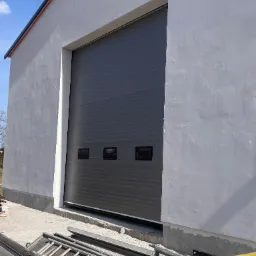 Szara brama segmentowa z trzema kwadratowymi okienkami, zamontowana w surowej elewacji budynku, widoczne drabiny i rury na ziemi.