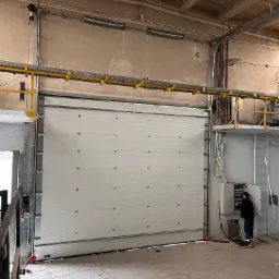 Biała segmentowa brama przemysłowa w hali, częściowo otwarta, z widocznym mechanizmem podwieszonym do sufitu; pracownik w kombinezonie obsługuje panel sterowania po prawej stronie.