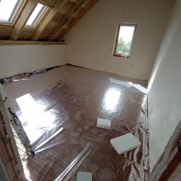 INSAJ Usługi hydrauliczne Poznań - Montaż ogrzewania podłogowego w nowym domu: widok na rozłożoną matę grzewczą z czerwonymi kablami na folii odbijającej ciepło, białe płyty izolacyjne styropianowe, surowe ściany i drewniana...