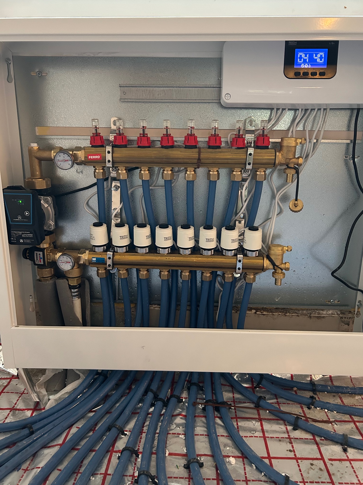 Rozdzielacz ogrzewania podłogowego z rurami w kolorze niebieskim, zamontowany w białej szafce, widoczny sterownik elektroniczny z wyświetlaczem i manometry.