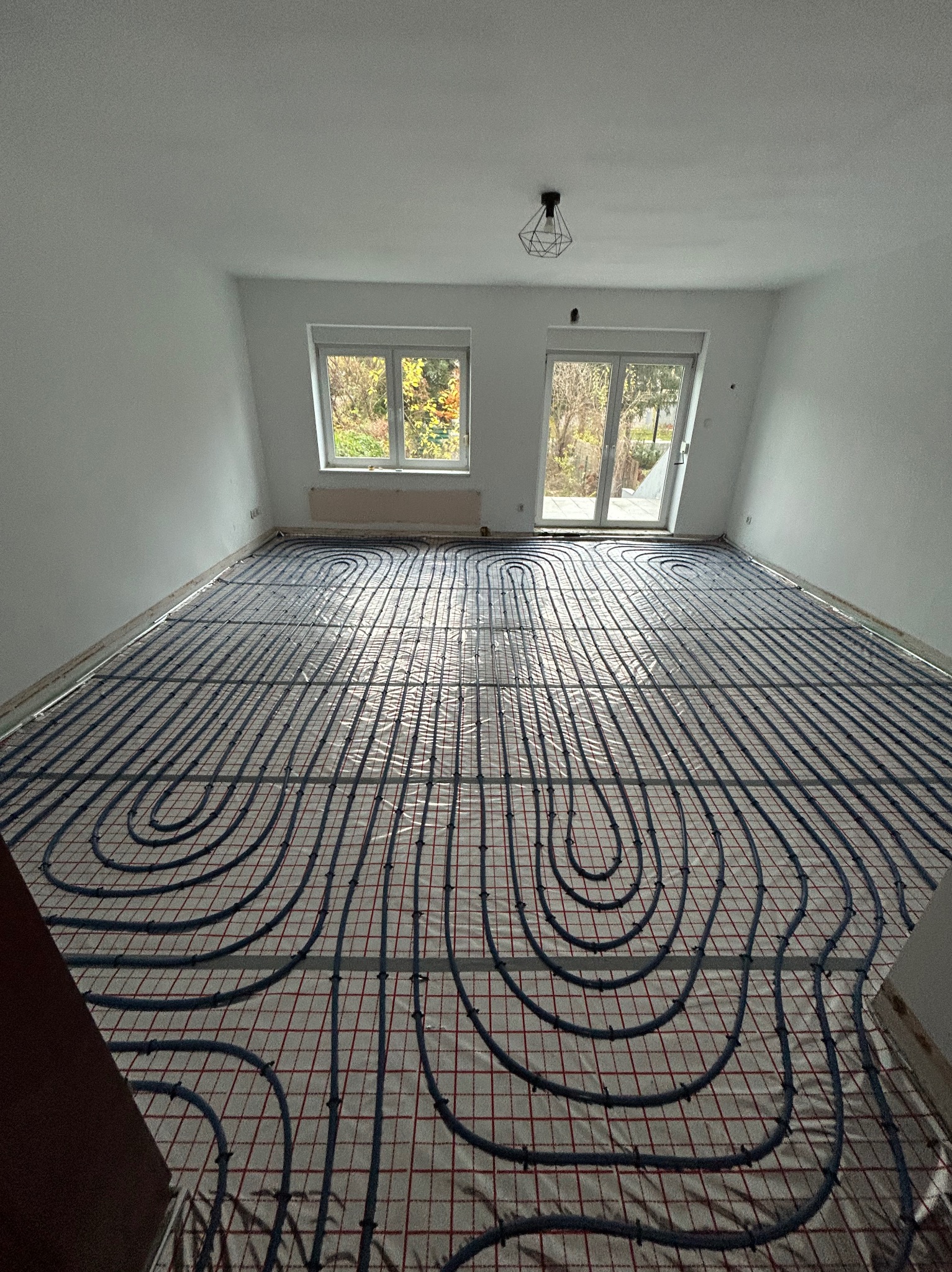 Rozległy pokój z białymi ścianami i oknami, w trakcie instalacji ogrzewania podłogowego, widoczne rury ułożone w pętle na siatce z czerwonymi liniami.