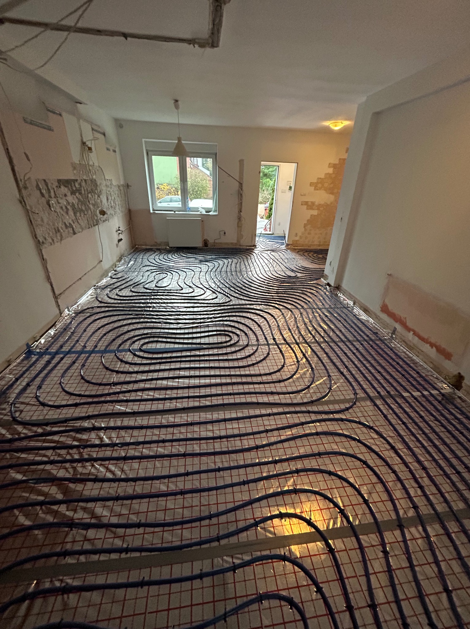 Niebieskie rury ogrzewania podłogowego ułożone na folii odblaskowej w trakcie instalacji w remontowanym pomieszczeniu z odsłoniętymi fragmentami ścian.