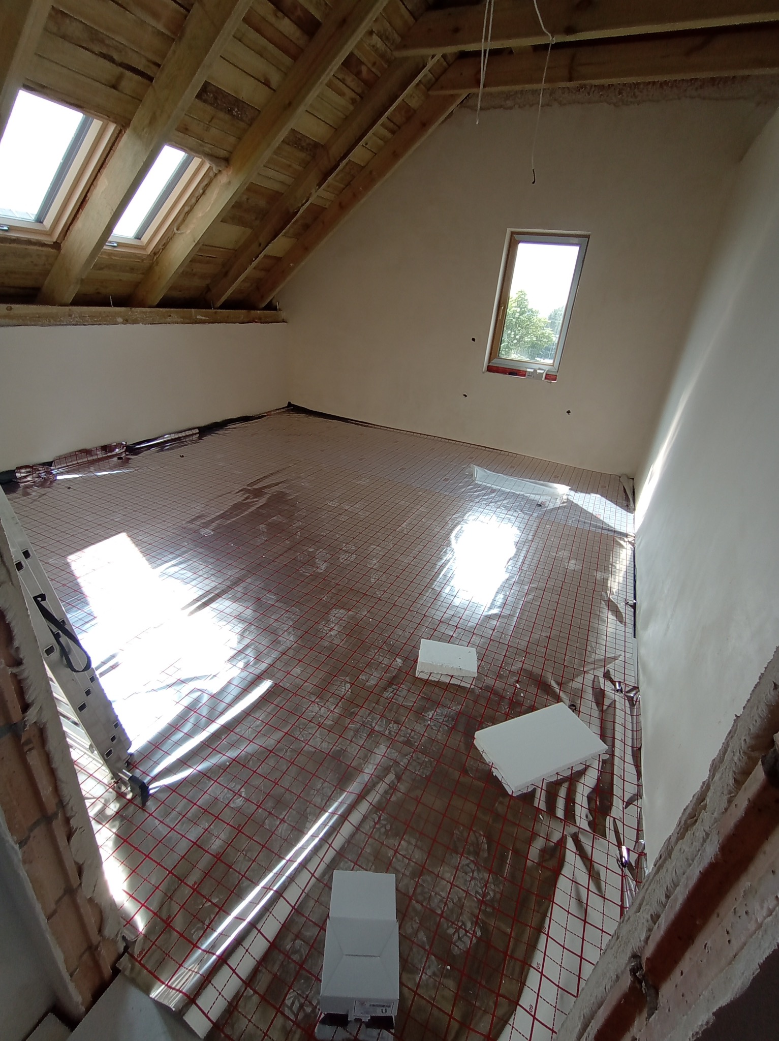 Montaż ogrzewania podłogowego w nowym domu: widok na rozłożoną matę grzewczą z czerwonymi kablami na folii odbijającej ciepło, białe płyty izolacyjne styropianowe, surowe ściany i drewniana...