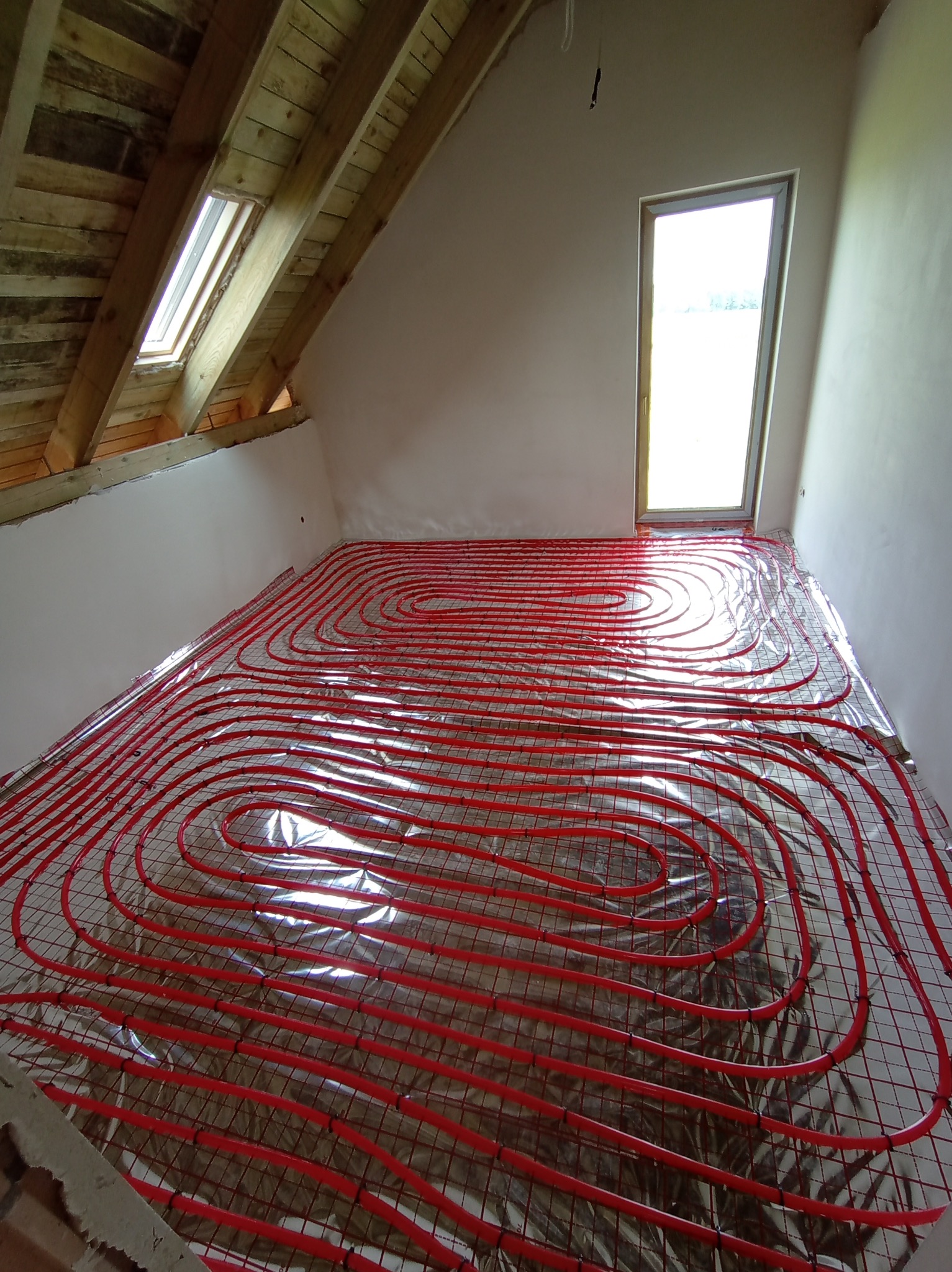 Czerwone rurki ogrzewania podłogowego ułożone na srebrnej folii refleksyjnej, widoczne drewniane belki stropowe i okno dachowe w surowym stanie.