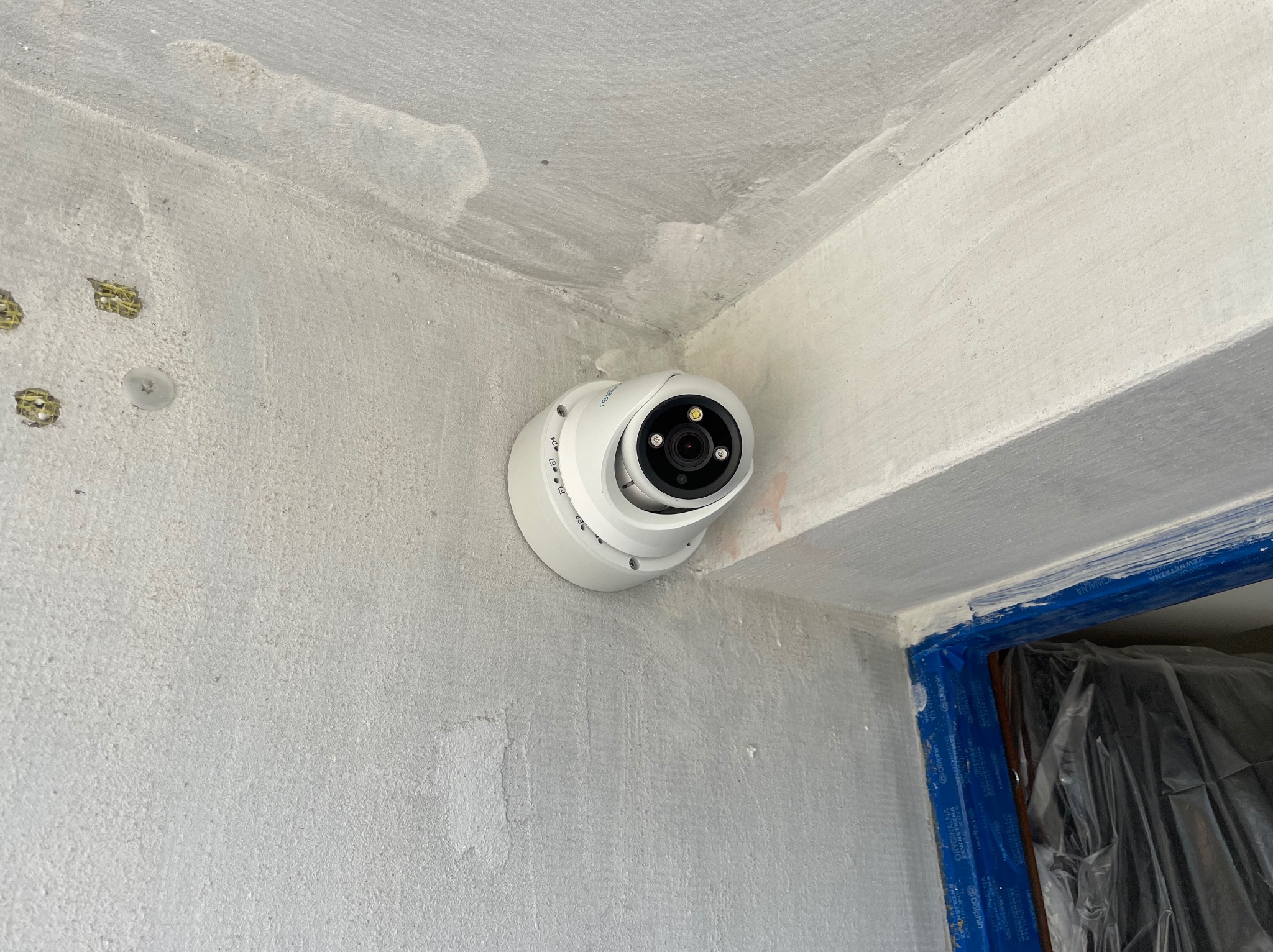 Biała kamera kopułkowa zamontowana w rogu pomieszczenia na surowej, szarej ścianie, widoczne ślady gładzi i zabezpieczone folią malarską ościeżnice.