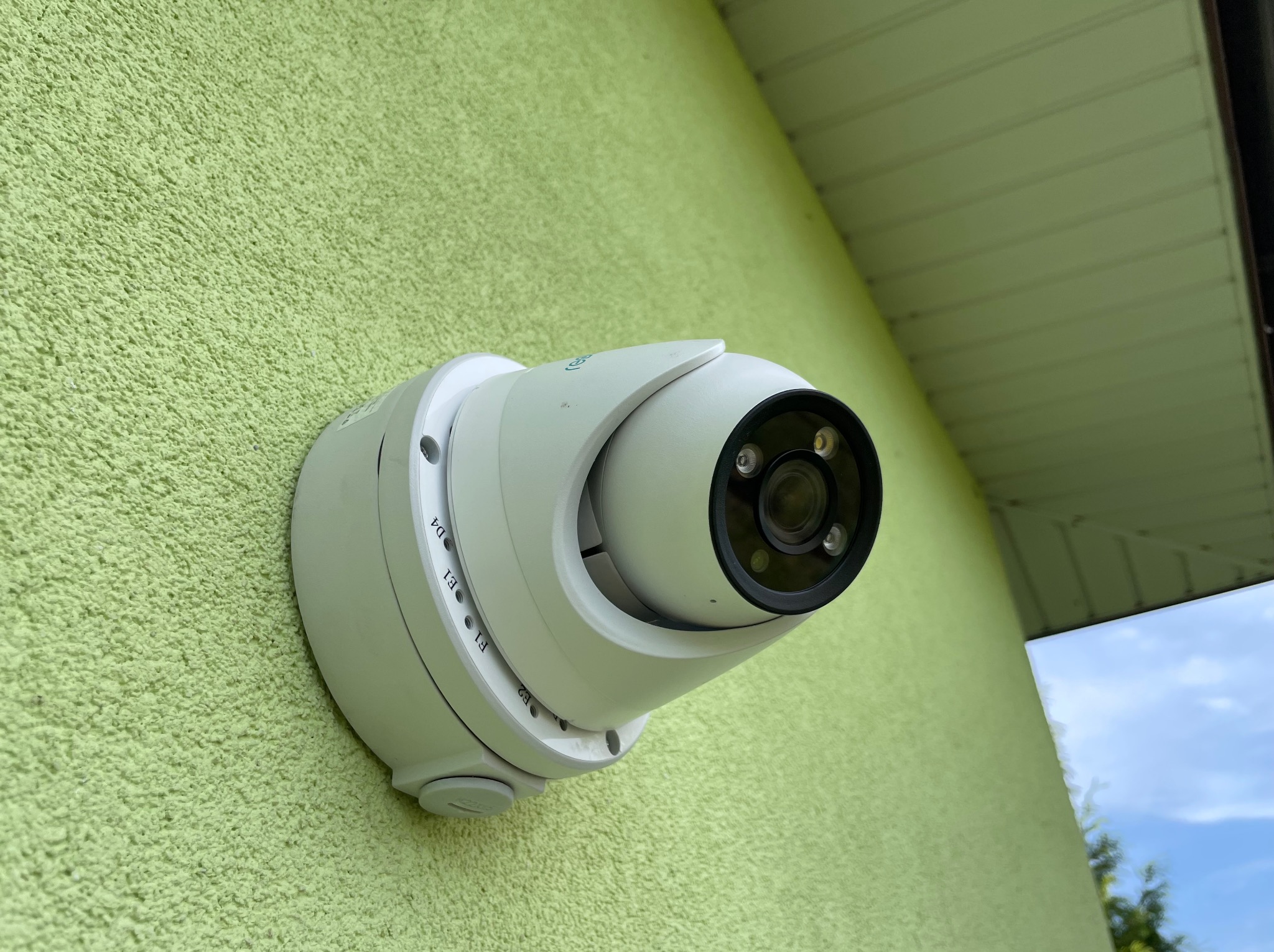 Zewnętrzna kamera obrotowa IP zamontowana na zielonej, teksturowanej ścianie budynku, widok z dołu.