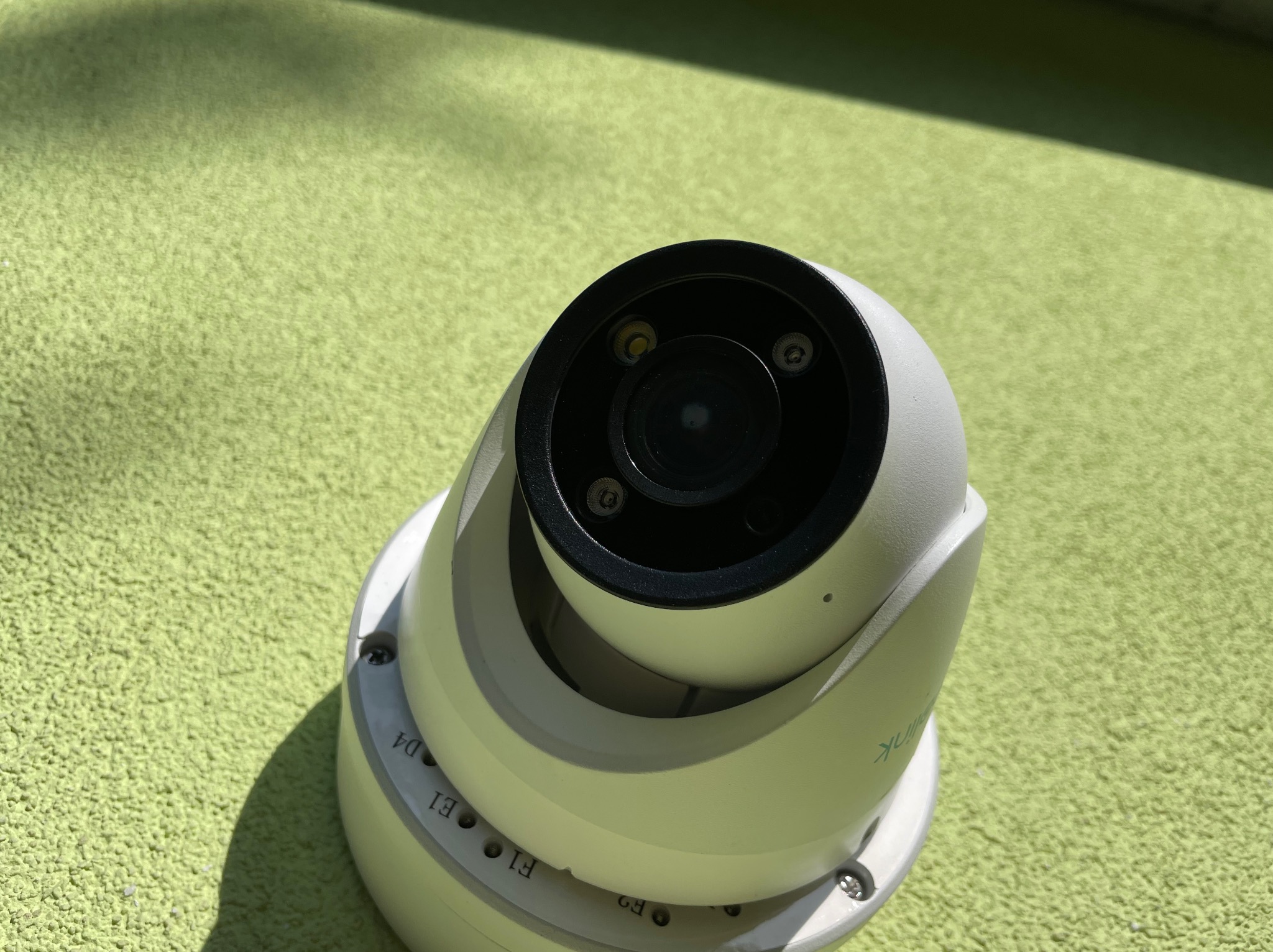 Kamera kopułkowa CCTV Reolink, widok z góry na zielonym tle o szorstkiej fakturze, oświetlona bocznym światłem słonecznym.