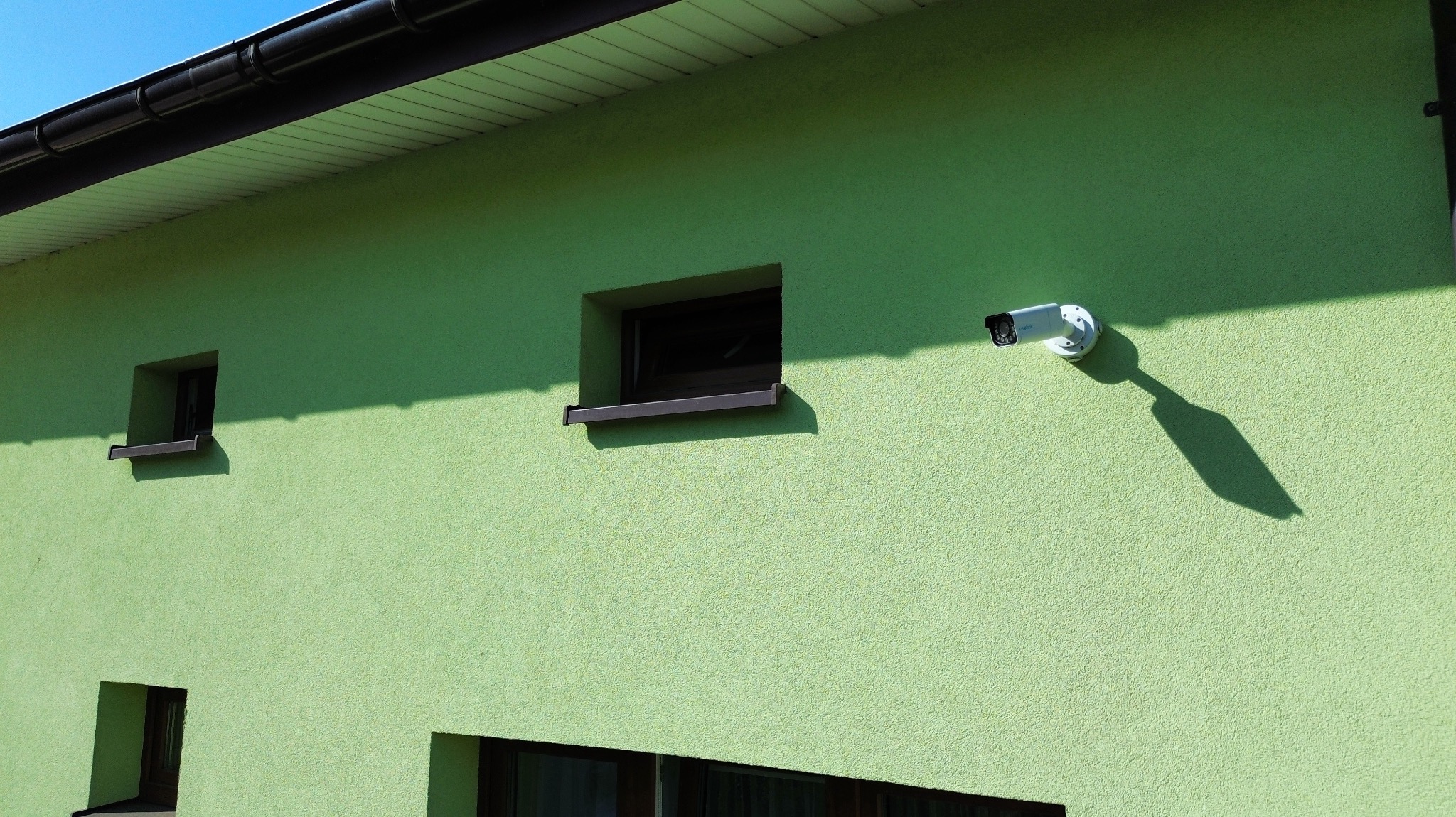 Zewnętrzna kamera monitoringu zamontowana na ścianie domu o jasnozielonej elewacji, widoczne okna z ciemnymi ramami i fragment dachu.