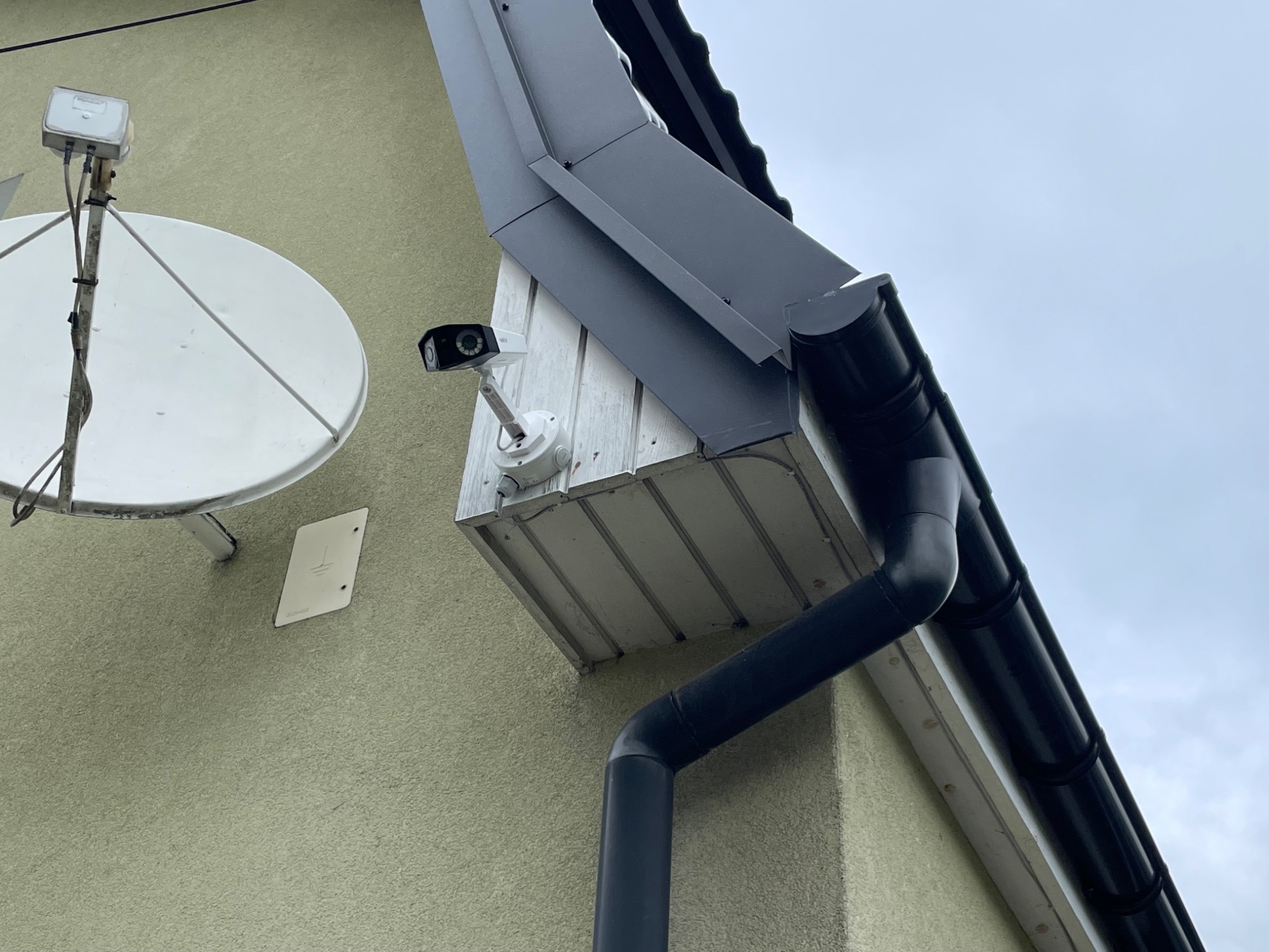 Zewnętrzna kamera monitoringu zamontowana na elewacji domu obok rynny i anteny satelitarnej.