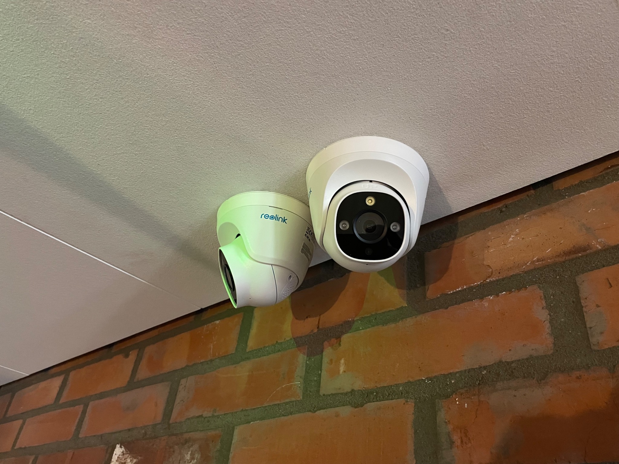 Dwie kamery Reolink zamontowane pod sufitem, z widoczną ścianą z czerwonej cegły poniżej.