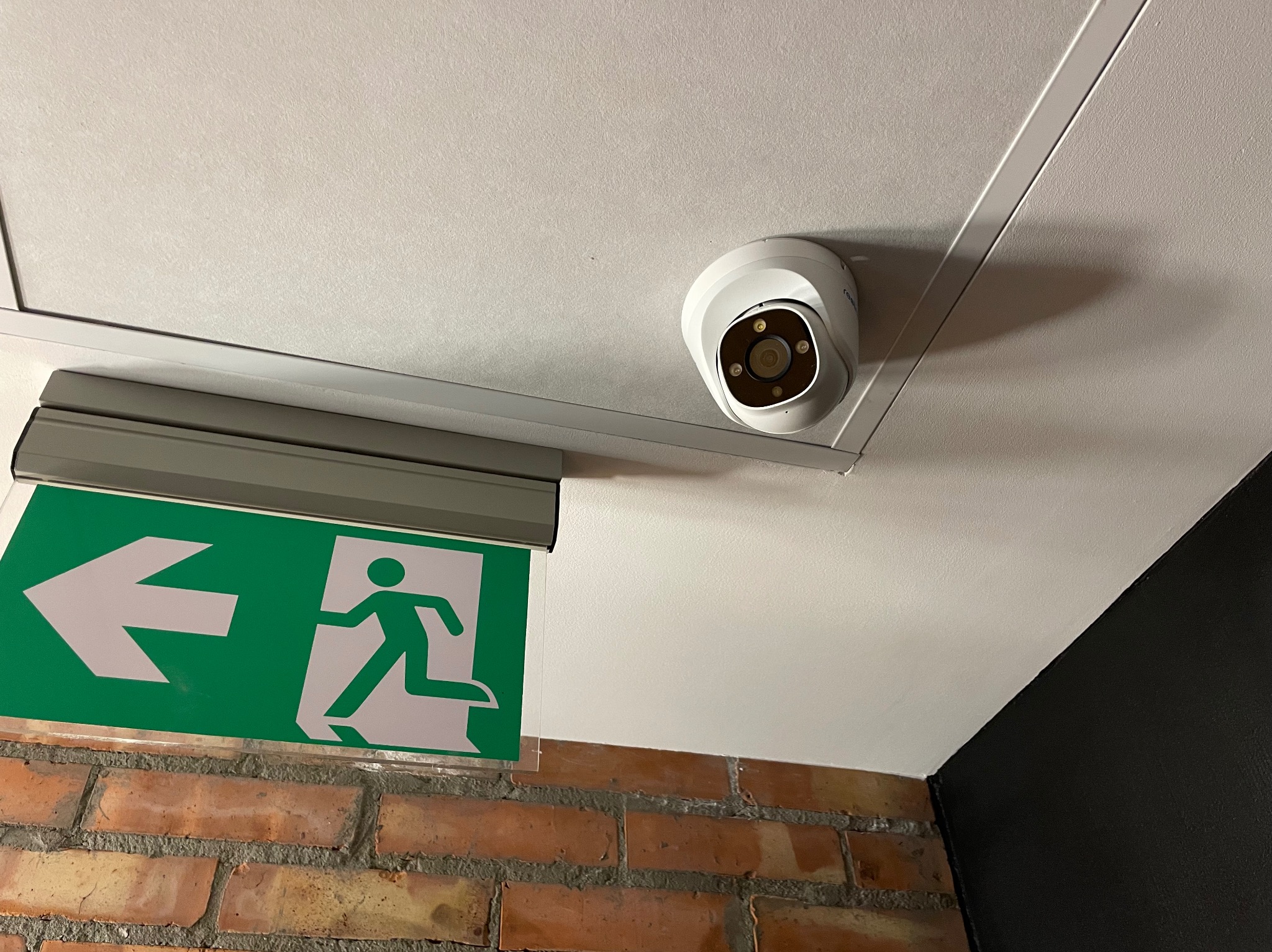 Biała kamera kopułkowa zamontowana na suficie obok zielonego znaku ewakuacyjnego z białym piktogramem biegnącej osoby, widok z dołu na ceglaną ścianę.