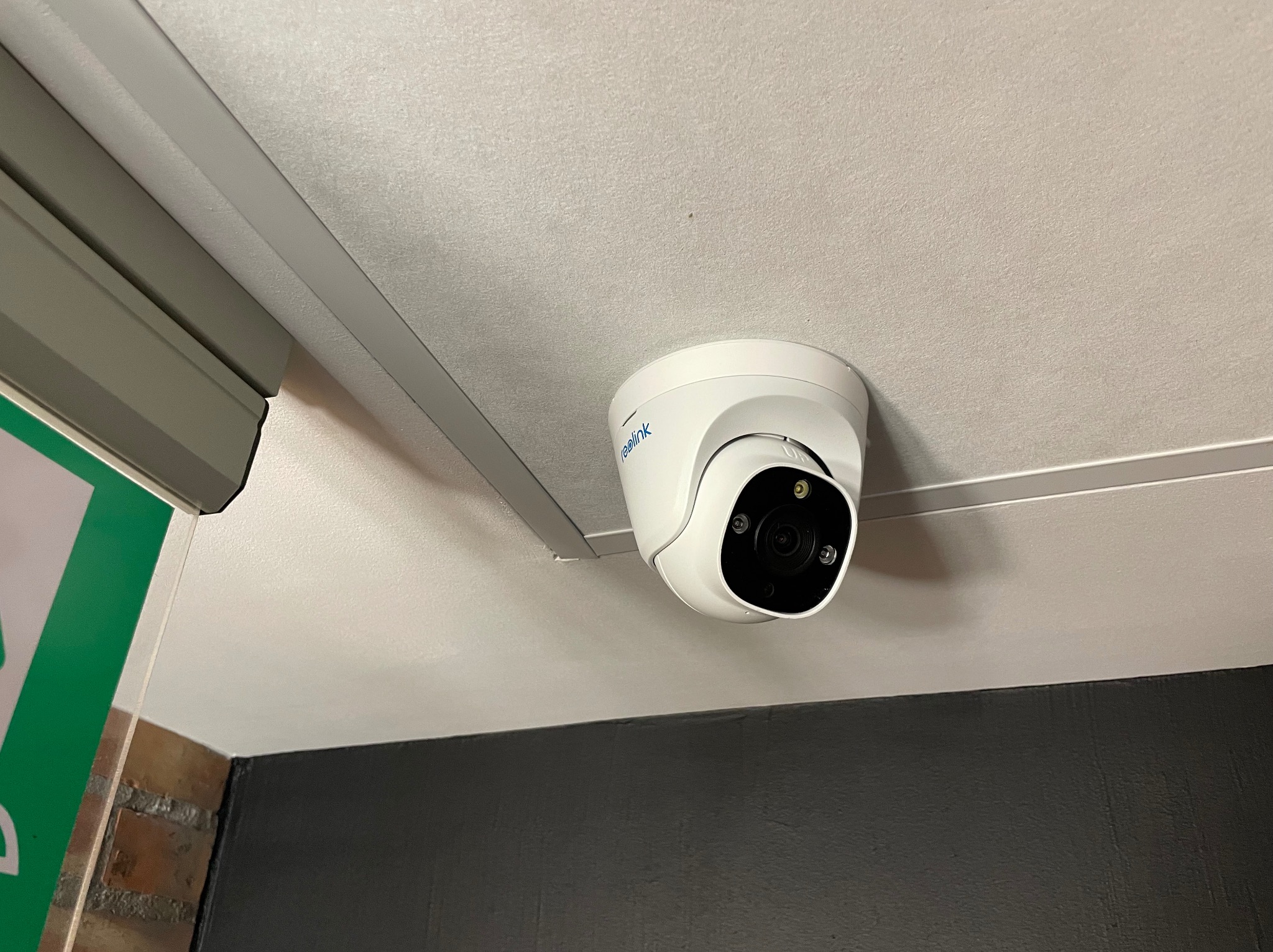 Biała kamera Reolink kopułkowa zamontowana na suficie, widoczny fragment ściany pomalowanej na czarno i fragment ramy okiennej z roletą.
