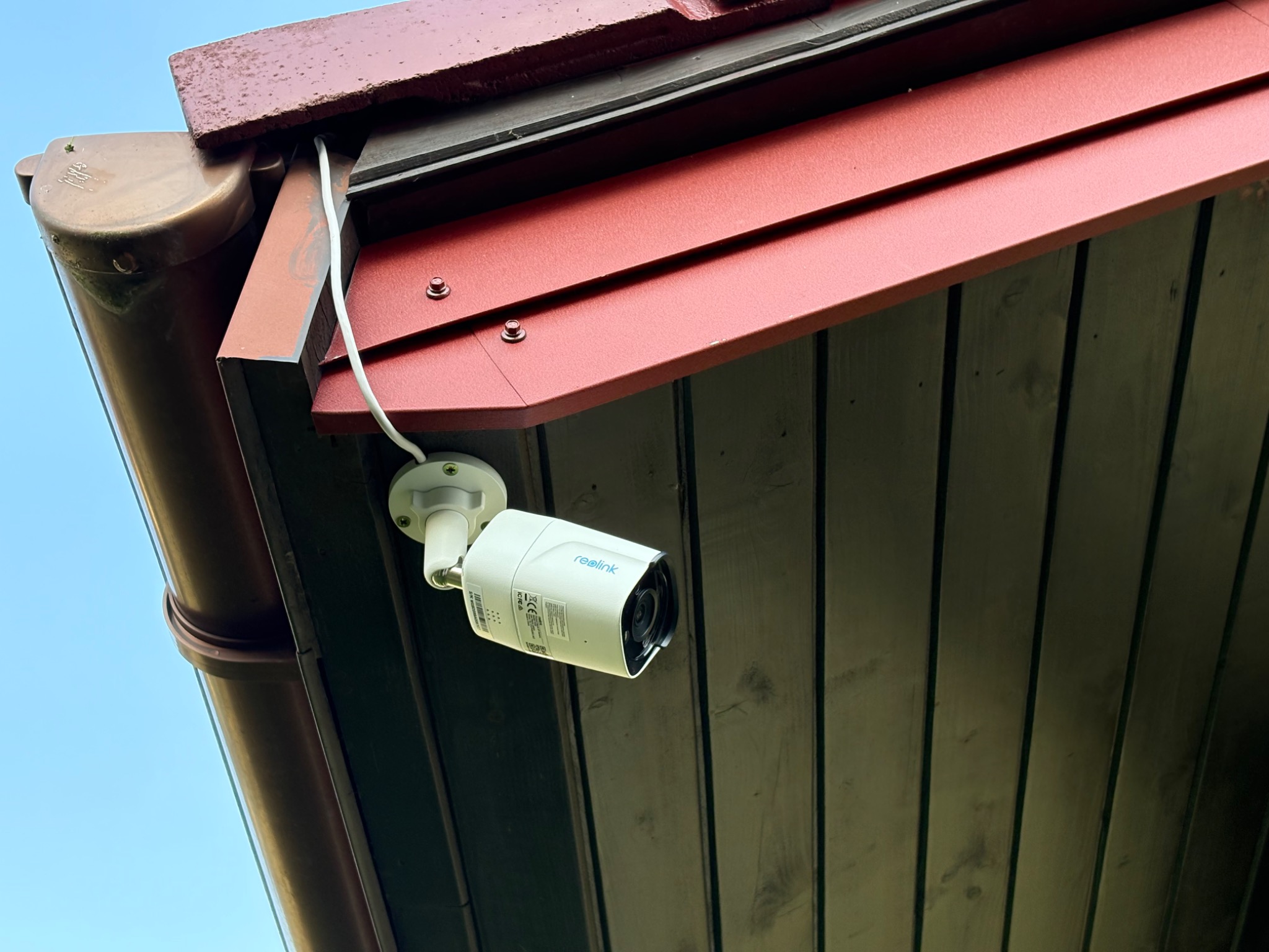Kamera Reolink zamontowana pod okapem dachu z brązową rynną, widok z dołu na błękitne niebo. Widoczne przewody i elementy mocujące.