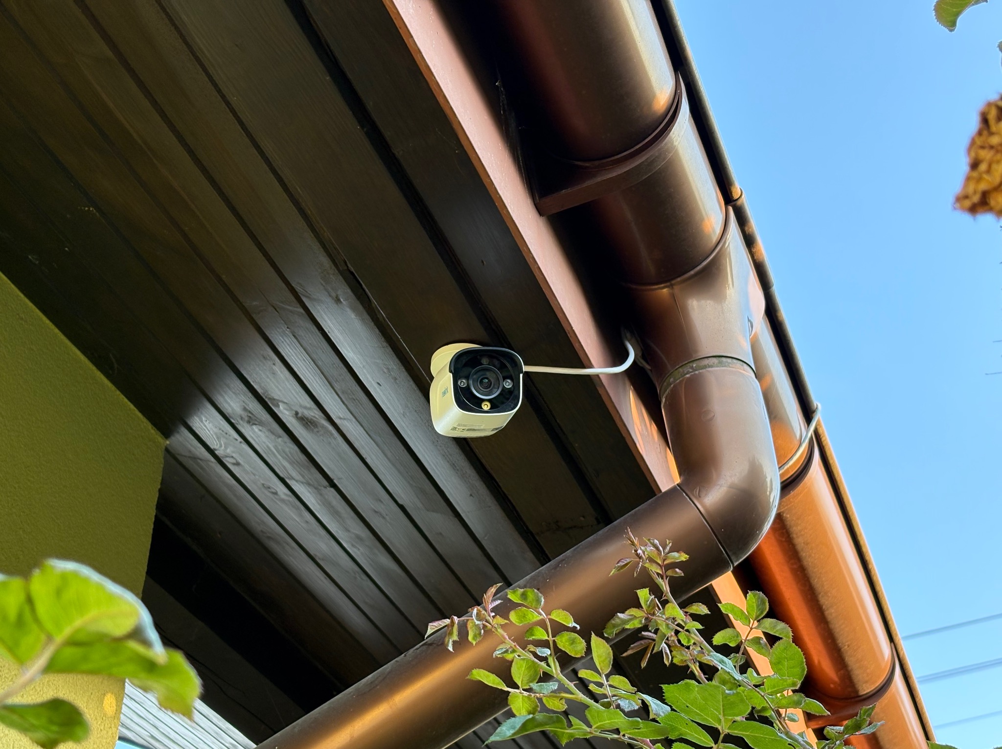 Kamera monitoringu przymocowana do drewnianej podbitki dachu obok rynny. Widok z dołu, na tle błękitnego nieba. Widoczne fragmenty roślinności.