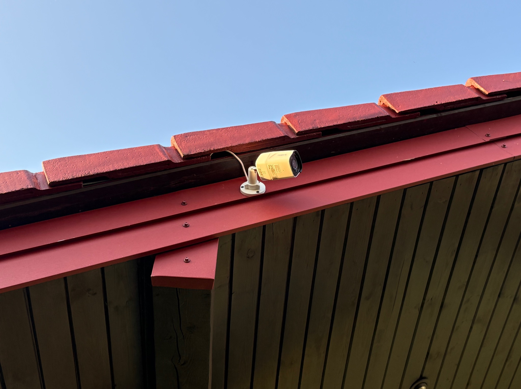 Kamera monitoringu Reolink zamontowana na czerwonym dachu drewnianego domu, widok z dołu na błękitne niebo. Instalacja systemu zabezpieczeń.