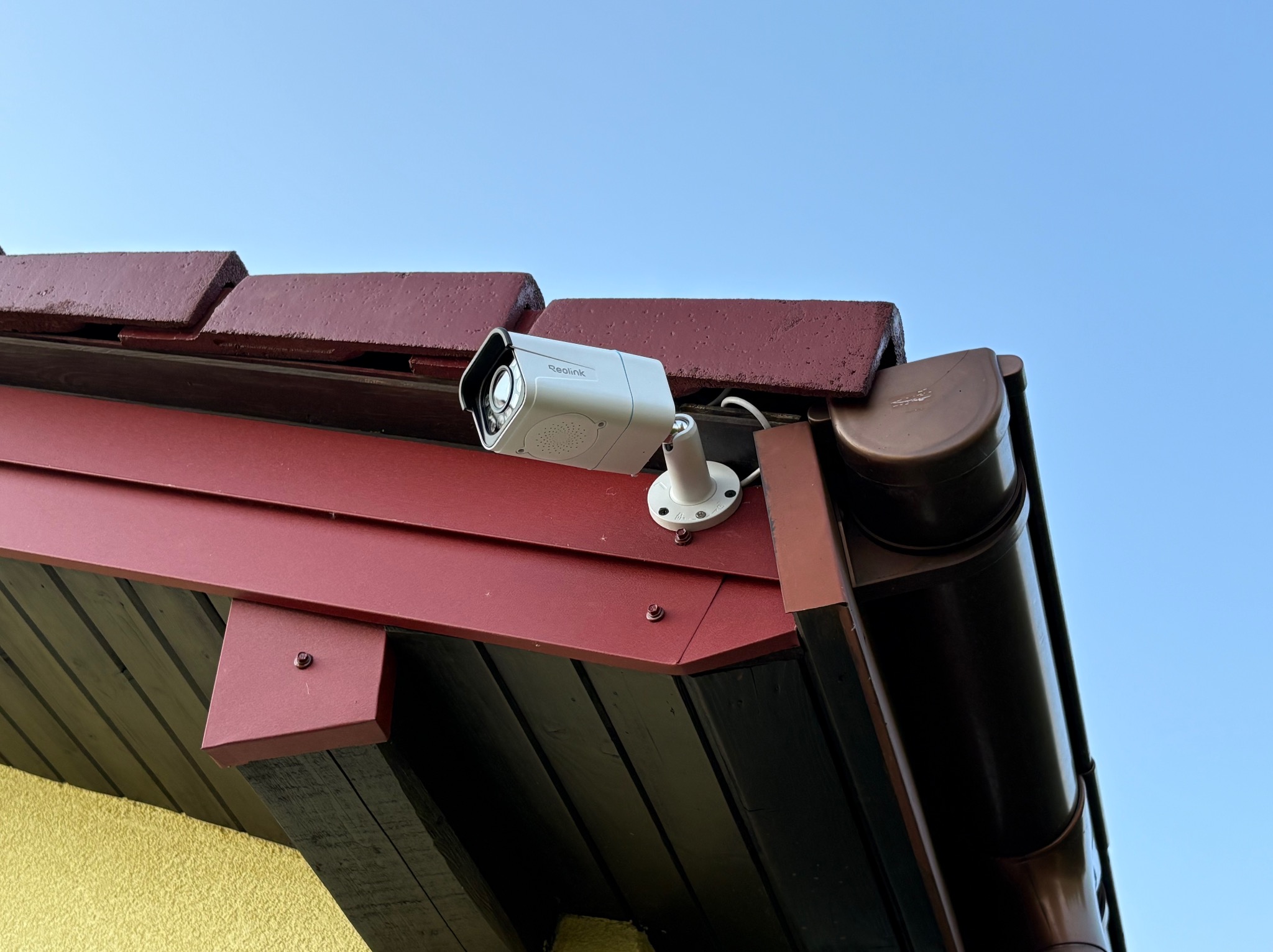 Kamera Reolink zamontowana na czerwonym dachu domu, z widocznym brązowym systemem rynnowym i fragmentem żółtej ściany. Błękitne niebo w tle.