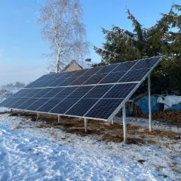 Panel słoneczny na metalowej konstrukcji, pokryty śniegiem, na tle zimowego krajobrazu z drzewami i częściowo widocznym domem.