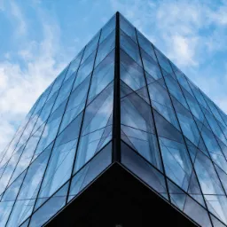 Szklany narożnik nowoczesnego biurowca odbijający błękitne niebo z chmurami.