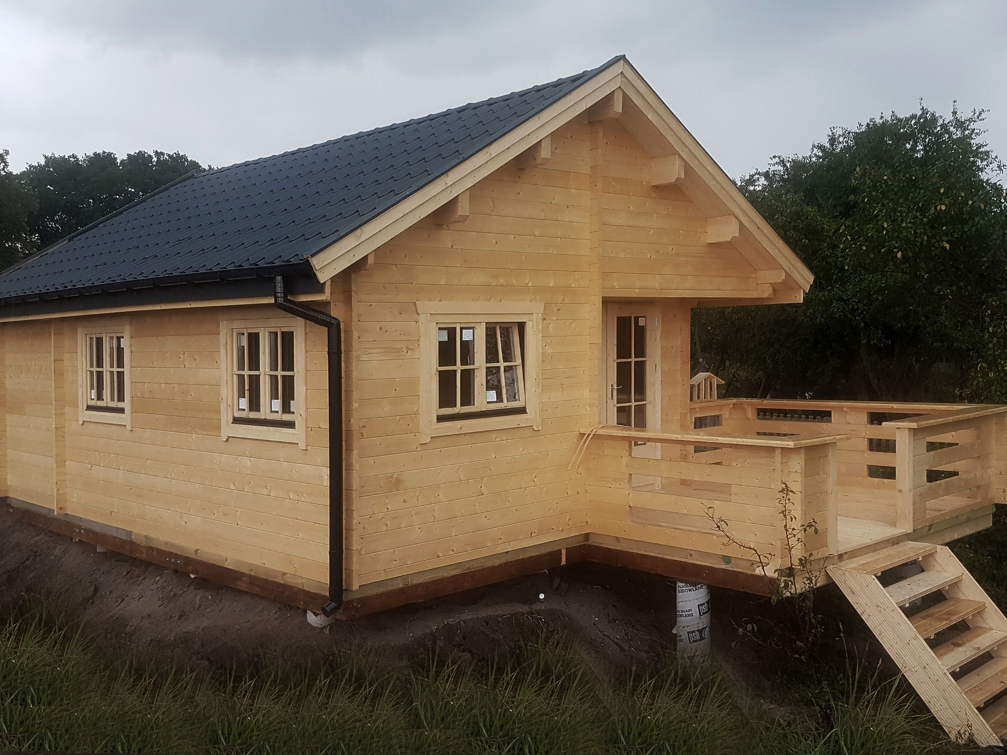 Surowy, drewniany domek letniskowy z ciemnym dachem i małym tarasem, widoczny z boku, na tle zieleni i pochmurnego nieba.