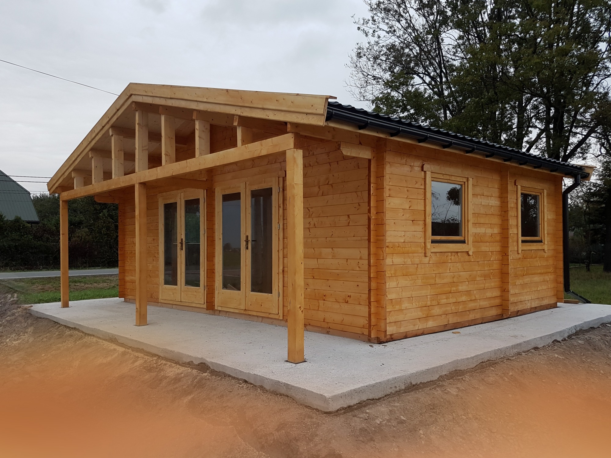 Drewniany domek letniskowy z przeszklonymi drzwiami i oknami, zadaszony ganek z odkrytą konstrukcją więźby dachowej, posadowiony na betonowej wylewce.