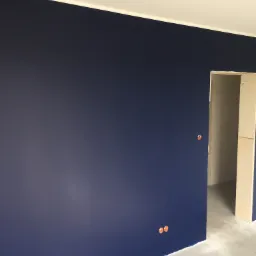 Świeżo pomalowana ściana w odcieniu głębokiego granatu z widocznymi otworami na gniazdka elektryczne i fragmentem surowej podłogi z kartonami ochronnymi.