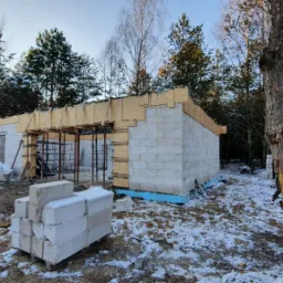 Budowa domu w stanie surowym otwartym zimą, widoczne bloczki Ytong, drewniane szalunki, rusztowania i ośnieżone drzewa w tle, słońce przedziera się przez gałęzie.